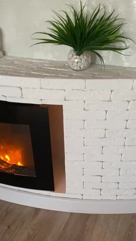 The video pans across a white brick electric fireplace. The flames within the fireplace are lit, displaying an orange glow. On top of the fireplace, there is a white statue of two figures embracing, a vase with green faux foliage, and a collection of white pebbles. The fireplace is situated on a light-colored wooden floor. The background wall has a textured, light gray finish.