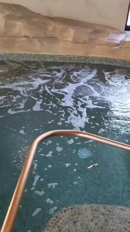 A brief video shows a tiered, jacuzzi-style pool indoors. The pool is tiled with small, square, teal tiles and surrounded by faux-stone material. A copper railing runs around the pool's edge and down the stairs leading into it. Bubbles rise from the water, indicating jets are on. A white column is in the middle of the pool area, and a window is visible above the pool. The camera pans slightly to show the different levels of the pool.