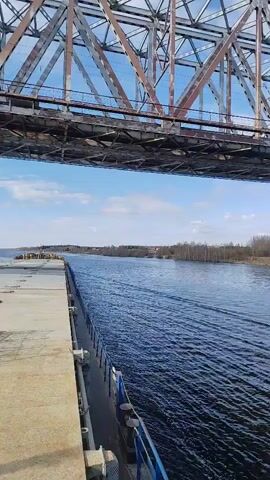 The video shows a view from a boat passing under a steel truss bridge over a body of water. The river water is dark blue and rippled. On the right bank, there's a flat grassy area with bare trees and a few buildings visible in the distance. The bridge is a complex structure of riveted steel beams with some rust visible. The bridge supports are concrete. The sky is light blue with scattered white clouds.