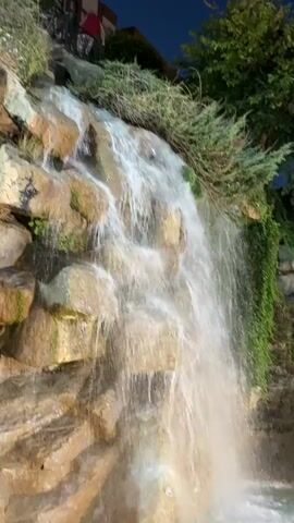 A man-made waterfall cascades down a rocky incline covered with greenery. The water flows over tan and brown rocks, creating a frothy white effect at the bottom where it enters a pool of turquoise water. The surrounding environment appears to be outdoors at dusk, with artificial lighting illuminating the scene. A hint of a building or structure can be seen in the background.