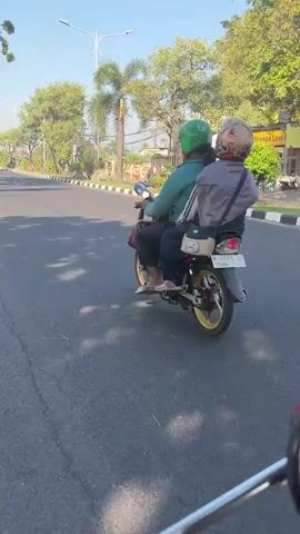 The video shows a street scene with a clear blue sky and sunlight. The camera is mounted at a low angle, likely on a motorcycle, as indicated by the partial view of handlebars and a mirror in the foreground. The video begins by focusing on two people riding a motorcycle. The person in front is wearing a green helmet, and the person behind is wearing a red and white helmet and a gray jacket. They are riding away from the camera.

The camera then pans slightly to the right, revealing a white truck carrying multiple large blue water jugs stacked on its flatbed. The truck is traveling in the same direction as the motorcycle, to the right of the frame. The road appears to be asphalt with a white dashed line marking the lane. On the left side of the road, there are trees, a sidewalk, and some buildings, one of which has a sign that reads "MR. DAY Always Love". On the right side of the road, there are more trees and plants. The overall environment suggests a sunny day in an urban or suburban setting.