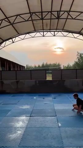 In a semi-outdoor training area with a metal roof and concrete walls, several young people are engaged in physical activity. The ground is covered with blue interlocking mats, some of which appear wet. In the background, a fence separates the training area from a grassy field with trees and buildings. The sky suggests it's late afternoon or early evening, with soft, warm lighting. The overall mood is one of active training and camaraderie.

The subjects include multiple individuals, predominantly young males, in various stages of a physical exercise or martial arts practice. They are dressed in athletic wear, including t-shirts, shorts, and what appears to be a gi. Some are in a crouched position, while others are in motion.

The activity appears to be a form of athletic training, possibly martial arts or calisthenics, involving dynamic movements and bodyweight exercises.

Notable details include the overcast but warm sky, indicating it might be nearing sunset. The wet mats suggest recent rain or a cleaning. The presence of buildings and trees in the background hints at a rural or semi-rural setting, consistent with the provided location context of Krasnoturansk, Russia. There is no visible text in the media file.