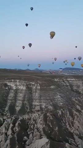 A drone footage showcases a vast, arid landscape with dramatic rock formations in the foreground. The sky is a clear, pale blue, transitioning to a soft pink and orange hue near the horizon, indicating either sunrise or sunset. Numerous hot air balloons, of various colors including white, yellow, and patterned designs, are scattered across the sky at different altitudes. The balloons are ascending, appearing as small dots against the expansive backdrop. Below the balloons, a distant town or settlement is visible, nestled among rolling hills and further rock formations. The overall impression is one of a breathtaking aerial spectacle in a unique geological setting.