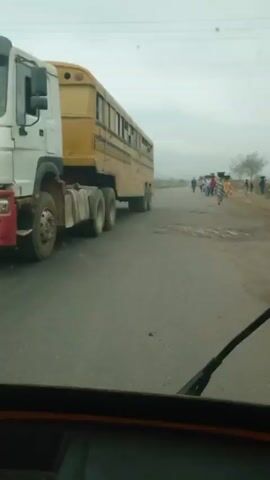 The video shows a view from inside a vehicle driving down a road. A yellow bus is to the left of the vehicle, and a group of people are walking on the right side of the road. Further down the road, a tanker truck is visible. The sky is overcast. The road surface appears to have some potholes. The vehicle's windshield wiper is visible in the foreground.