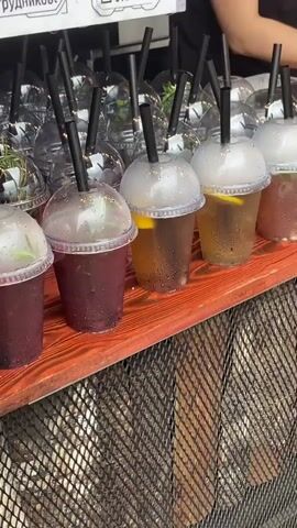 The video shows a close-up of a row of drinks with black straws in plastic cups. The drinks are of various colors, including purple, amber, and reddish-brown. Some of the cups have condensation on them, suggesting they are cold. There are also slices of lemon and sprigs of herbs visible in some of the drinks. The cups are arranged on a wooden surface that appears to be part of a food stall or counter. In the background, more drinks in plastic cups are visible, along with some blurred figures of people. The overall impression is of a refreshing beverage stand.
