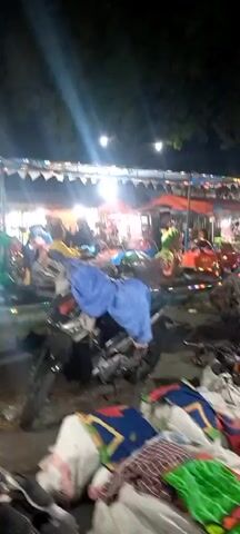 THE VIDEO SHOWS AN OUTDOOR MARKET AT NIGHT. PEOPLE ARE WALKING AROUND AND SHOPPING. THERE ARE SEVERAL STALLS SELLING CLOTHING AND OTHER GOODS. THERE ARE ALSO SEVERAL MOTORCYCLES PARKED IN THE AREA. THE MARKET IS ILLUMINATED BY BRIGHT LIGHTS.