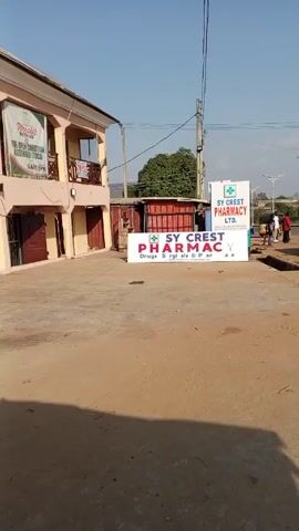 The video shows a street view during the daytime, focusing on an area with several signs. A building on the left displays a sign for "WORSHIP WITH US - THE OPEN CHRISTIAN ASSEMBLY (TOCA) - 9AM-1PM." In front of it is a dirt or concrete lot, leading to a road. Two signs for "SY CREST PHARMACY LTD." are visible. On the right side of the road, people are walking, and a silver car drives past. There are street lights along the road, and some trees in the background. The sky is clear and blue. The ground is dusty and uneven, with visible drainage systems along the road.