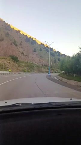 The video shows a view from inside a vehicle looking out at a highway curving through a mountainous, forested area. The sun appears to be low in the sky, casting a warm light on the hills. There are streetlights lining the road, and a truck can be seen in the distance on the highway. The road is paved and has lane markings. The scenery is dominated by green trees and brown, rocky hillsides.