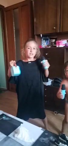 A young girl with blonde hair, wearing an oversized black t-shirt, stands in a room with wooden floors and walls. She holds a light blue toy in her left hand and a light blue microphone in her right hand, which she appears to be speaking into. Her mouth is open, and she is looking towards the camera with a joyful expression. Her body is angled slightly to her right, and her right arm is extended. In the background, there is a wooden cabinet with shelves holding various items, and a wooden door on the left. Another child, a boy with dark hair, is partially visible on the right side of the frame, looking down and holding something in his hands. A partially visible water bottle is in the foreground. The scene is likely indoors, possibly a home. The girl is engaged in what appears to be a playful activity, possibly singing or talking into the toy microphone.