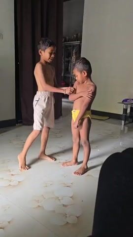Two young boys, likely brothers, are captured in a candid moment within what appears to be a residential setting in Nga, Indonesia. The older boy, standing on the left, wears light-colored shorts and is shirtless. He has a playful expression, looking towards the younger boy. The younger boy, on the right, sports yellow underwear and is also shirtless. He gazes down with a slightly more serious or focused look.

The scene is indoors, with a tiled floor featuring a floral pattern. A dark brown curtain hangs in the background, and a piece of electronic equipment, possibly a television, is mounted on the wall. A tall, dark cabinet with shelves is visible behind the older boy, and a speaker system is situated to his left. To the right, a table with a glass top and a lamp are present. The lighting suggests it could be daytime, with natural light possibly coming from an unseen window.

The boys appear to be engaged in some sort of interaction, possibly playing or preparing to move. The older boy seems to be reaching out or gesturing towards the younger boy. In the latter part of the clip, both boys are walking, with the younger boy moving towards an open doorway. A blue inflatable toy resembling a horse is visible in the background near the doorway, adding a touch of domesticity to the scene. The overall mood seems lighthearted and everyday. There is no visible text in the media file.