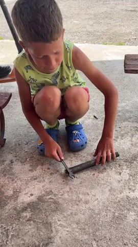 A young boy with short, dark hair is crouching on a concrete surface. He is wearing a light green tank top with a zebra pattern and red shorts. On his feet, he has blue Crocs. In his right hand, he holds a thin metal rod, which he is using to poke at a dark, cylindrical object lying on the ground in front of him. The object appears to be a piece of metal pipe. The boy looks down at the object for a moment, then looks up at the camera with a neutral expression. The background suggests an outdoor setting with a gravel or dirt ground and hints of wooden furniture. The sound accompanying the video is the distinct metallic clinking of the rod striking the pipe.