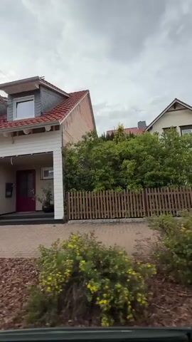 The video pans across the front of a white, two-story house with a red tile roof. The house has dormer windows on the upper level and a single window with a flower box on the lower level. The house number, 1175, is visible above the dark red front door. To the left of the house, another house with a dark gray facade is partially visible, separated by a fence. The ground in front of the houses is paved with bricks. The sky is overcast.
