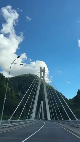 The video shows a first-person perspective of driving across a cable-stayed bridge. The bridge has two tall, white, A-frame pylons supporting numerous cables that stretch to the road surface. The road is asphalt with white lane markings. The bridge spans across a valley with lush green mountains on either side, covered in trees. The sky is a bright blue with scattered white clouds. There is a street lamp visible on the left side of the bridge. The camera appears to be mounted inside a vehicle, looking forward. The movement of the camera suggests the vehicle is in motion, driving towards the center of the bridge.