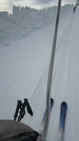 The video depicts a first-person perspective of someone riding a ski lift up a snowy slope. We can see a portion of the lift's metal pole in the center of the frame. To the left are ski poles strapped to a grey backpack. Below the pole are the rider’s blue and black skis, moving steadily upward on the snowy ground. Tire tracks are visible in the snow. In the background is a large pile of snow, possibly snow-making remnants, with a few trees silhouetted against the overcast sky. There are other individuals on skis ahead and to the right, also riding the lift. The overall impression is of a calm, wintry scene at a ski resort.