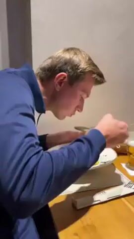 A man is shown eating a bowl of soup at a wooden table in a restaurant. He is wearing a blue and white long sleeve shirt. After taking a bite, he picks up his smartphone and takes a photo of the soup. Other items visible on the table include a basket of bread, a glass of beer, and silverware. The walls of the restaurant are plain.