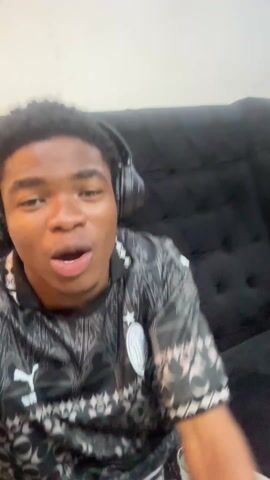 Two young men are sitting on a black couch. The man in the foreground is wearing a black and gray patterned shirt, headphones, and has short curly black hair. He is looking directly at the camera, smiling and gesturing with his hands. The man in the background is wearing a black t-shirt and khaki pants. He's seated with a device in his hands, looking upward and pointing. The background includes a wall with marble-like patterns, and an orange device. Upbeat music plays throughout.