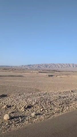 The video shows a wide, flat, arid landscape under a clear blue sky. In the foreground, there is a rocky roadside, transitioning into a flat expanse of dry land that stretches towards a distant range of mountains. Scattered across the flat area are a few small, seemingly abandoned, low-walled structures or ruins. The camera appears to be moving along a road, as indicated by the paved surface in the immediate foreground. The overall scene conveys a sense of sparseness and desolation.