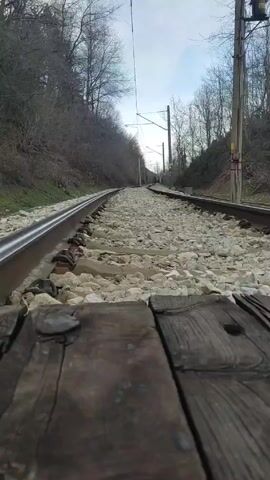 The video shows a ground-level view of a railway track. The tracks are flanked by a bed of grey ballast. On either side of the tracks, there are slopes covered with trees and some green vegetation. Overhead electrical lines are visible, supported by utility poles. The sky is overcast. The camera angle is pointed down the length of the tracks, creating a vanishing point in the distance.