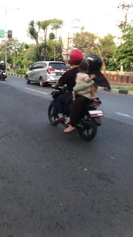 The video shows a street scene with several vehicles. In the foreground, a person wearing a blue helmet and a light-colored hooded sweatshirt is riding a scooter. They are wearing dark pants and appear to be moving away from the camera. In the middle ground, a black pickup truck with a cage-like structure in its bed is traveling in the same direction as the scooter. Further in the background, another scooter is visible, as is a white car. The environment appears to be a city street with trees and buildings lining the road. The lighting suggests it might be late afternoon or early evening. The overall impression is a moment captured from a moving vehicle, likely another motorcycle, observing the flow of traffic.