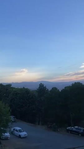 The video captures an outdoor scene with a clear blue sky fading into a pale horizon where distant mountains are visible. The foreground is dominated by lush green trees forming a dense canopy, with a winding road cutting through the landscape. Several cars are parked along the roadside. The overall atmosphere suggests a peaceful, natural environment, possibly during sunset or early morning, given the soft lighting.