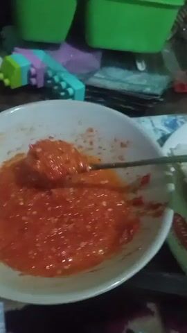 The video shows a close-up of a meal. There is a white plate with rice, a piece of fried chicken, and a spoon with red sauce being placed on the rice. There is also a white bowl containing more red sauce. In the background, there are blurry green containers and other objects on a table. The lighting is dim and the focus is on the food.