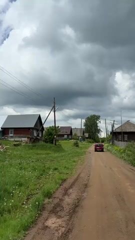 The video shows a rural scene on a cloudy day. A red car drives down a dirt road lined with traditional wooden houses. Utility poles and wires run along the road. Vegetation, including grass, weeds, and bushes, grows on either side of the road.  Puddles are visible in the road. Beyond the houses and vegetation, a forest is visible in the distance under a sky filled with dark, heavy clouds.