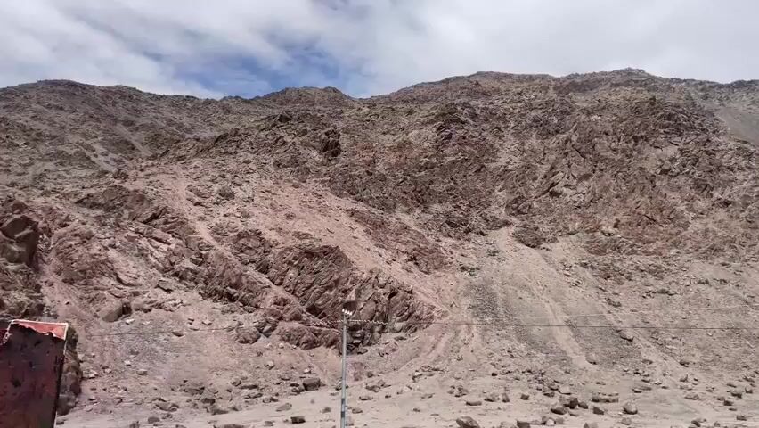 The video shows a panoramic view of a rocky mountain landscape under a cloudy sky. The mountains are steep and covered with loose rocks and scree, with prominent rock formations visible. In the foreground, there is a dirt road or track, and on the left side, a portion of a reddish structure, possibly a small building or shelter, is visible. A utility pole can be seen on the right side of the frame. The overall environment appears dry and arid, suggesting a mountainous region with limited vegetation. The camera appears to be moving horizontally, capturing the vastness of the mountainous terrain.