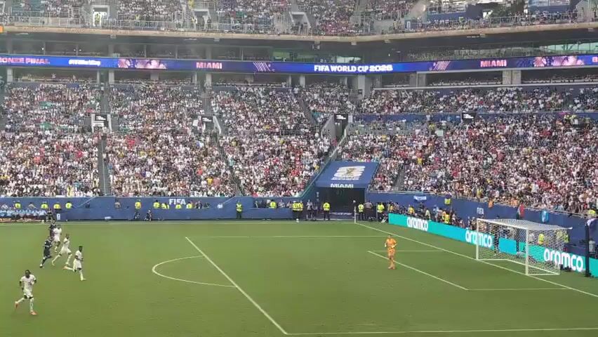 The video shows a soccer match being played in a large stadium filled with spectators. The game is in progress, with players from two teams on the field. One team is wearing dark blue uniforms, and the other team is wearing white uniforms. The ball is on the green grass field, near the center of the action. A goalkeeper in orange is positioned in front of the goal. In the background, a large crowd of people is seated in the stands, watching the game. Above the stands, signs display "MIAMI" and "FIFA WORLD CUP 2026." The environment appears to be daytime, with natural lighting.