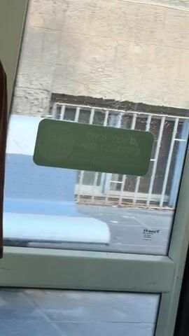 The video shows the interior of a light rail train. A young woman with dark braids is standing near a glass door. She is wearing a brown long-sleeved shirt with "NEW YORK" printed on the front and black pants. A green sticker is affixed to the glass door. The train is in motion, and the outside environment is visible through the glass. A silver handrail is visible near the door. The overall lighting is bright, suggesting daytime.