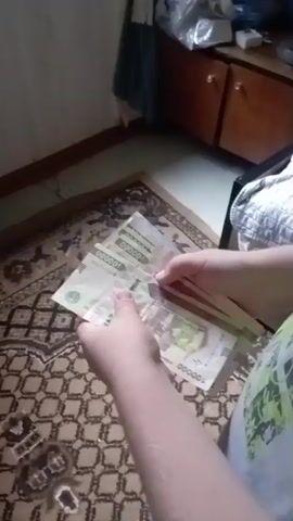 A person's hands are shown holding a stack of banknotes, the denominations visible are 100,000 and 50,000. The person is fanning the banknotes as if to count them. The background shows a patterned rug, a wooden cabinet, and a wall.