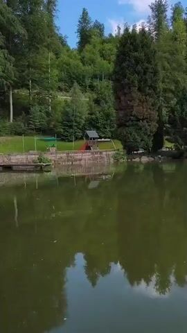 This media file is a video showing a scenic view of a lake surrounded by lush green trees and a hillside. The water is calm and reflects the trees and the sky, suggesting a clear day. On the edge of the lake, there is a small playground structure with a slide and a wooden hut, indicating it's a recreational area. A pathway or railing runs along the edge of the lake, separating it from the playground and the hillside. The trees are dense and varied, with some appearing to be pine trees, characteristic of a forest environment. The overall scene evokes a sense of tranquility and natural beauty. There is no visible text in the media file. The location is identified as Triberg im Schwarzwald, Germany, a region known for its natural beauty, waterfalls, and forests. The time of day appears to be daytime, given the brightness and the clear reflections in the water. The weather seems pleasant, likely sunny and warm, conducive to outdoor activities.