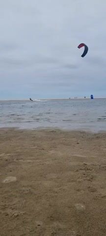 A wide shot of a beach scene shows a kite surfer in the distance. The kite surfer is on the water, being pulled by a large kite in the sky. The kite is red and blue and is clearly visible against the overcast sky. The water is calm, with small waves breaking on the shore. The beach is sandy and appears to be uncrowded. In the background, there are a few other people walking on the beach and another person kite surfing. The overall impression is one of a leisurely day at the beach, with a focus on the water sports.