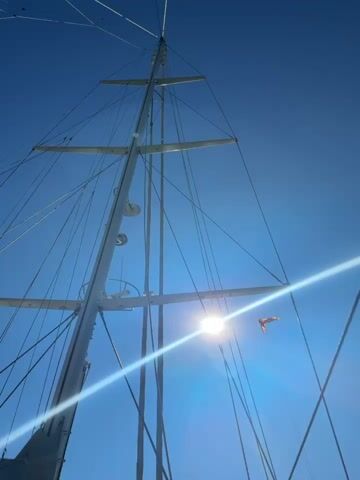The video shows a low-angle shot of the mast of a sailboat against a bright blue sky, with the sun shining through the rigging. A red flag flutters from one of the horizontal spreaders. The video then transitions to a wider shot of the calm, dark blue water surrounding the sailboat, with lush green mountains in the background. Finally, the video focuses on the wake of the boat, showing white foam against the deep blue water, with the shoreline reflected on the water's surface.