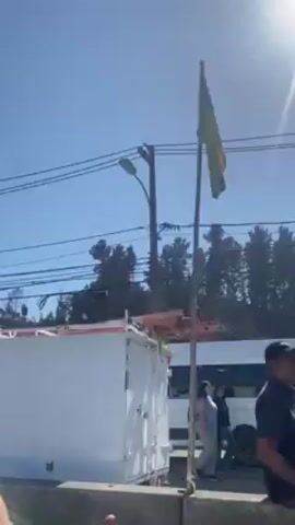The video, shot from a moving vehicle, shows a roadside view of a densely populated hillside. Many small structures, some of which appear to be temporary dwellings, are visible on the hillside, interspersed with trees. The Chilean flag is visible in several locations. Debris and construction materials are scattered around the hillside.  Several vehicles, including a white van and a truck carrying portable toilets, can be seen on the road.  Power lines and utility poles are visible overhead.  A sign that says "Se recibe ayuda" (Help is received) is partially visible. The sky is clear and blue.