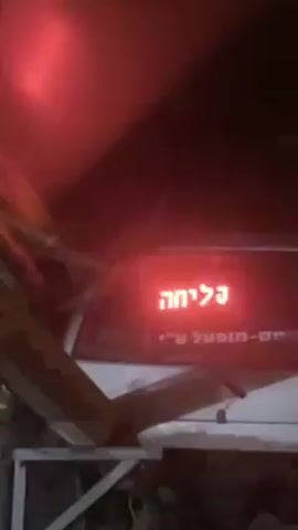 At night, the back of a white vehicle is illuminated by a red light. The Hebrew word "פליחה" which means "spitting" or "expectoration," is lit up in red on the back window. Below this word, a longer inscription in Hebrew is partially visible, which includes the words "מופע" and "עין." The camera then zooms out, showing a dark environment with structural elements in the foreground, but no people are visible in the scene.