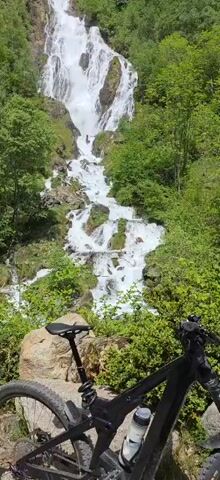 A high angle shot shows a waterfall cascading down a mountainside. The water is white and turbulent, and the rocks around the falls are covered in green moss. Lush green trees and bushes are visible on either side of the falls. In the foreground, the back of a black mountain bike is partially visible, resting against a rock and nestled in the greenery. The camera slowly zooms in on the waterfall. A few people are visible on the right side of the falls, sitting and enjoying the view.
