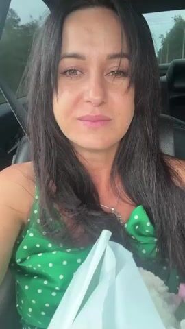 The media file is a short, first-person video of a woman holding a bouquet of flowers. She is in the driver's seat of a car, indicated by the car interior visible in the background. The bouquet is composed of white chrysanthemums and pink alstroemeria. The woman has long, dark hair and is smiling. The video appears to have been filmed during the day, with natural light filtering into the car. There is no visible text in the video. The setting is likely a road or parking area in Markova, Russia, given the provided location context. The woman's expression suggests she is happy or pleased, possibly having just received the flowers or on her way to a special occasion.