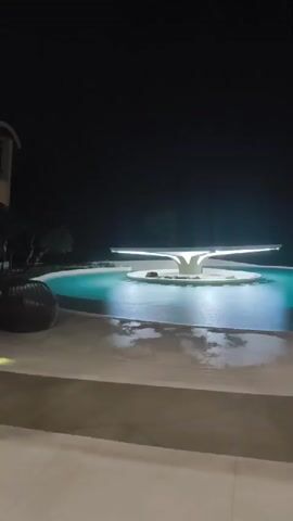 The video shows a nighttime view of an outdoor swimming pool. The pool is illuminated by underwater lights, casting a blue-green glow. In the center of the pool, there is a modern-looking fountain or sculpture with a flat, illuminated top and a white, sculptural base. The surrounding area appears to be paved with concrete or stone, and there is some foliage and a dark, shadowy background suggesting trees or landscaping. The overall impression is one of a luxurious and tranquil setting. The camera pans slowly across the scene, giving viewers a comprehensive look at the pool and its surroundings. The footage is shot from a slightly elevated perspective, looking down at the pool. There are no people visible in the video.