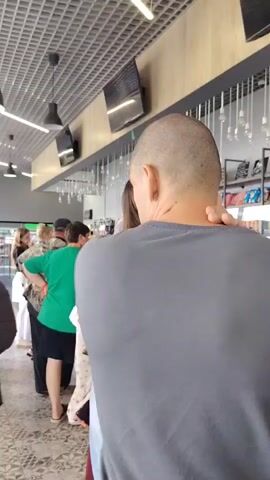 The video shows a line of people in what appears to be a store or cafe. The camera is focused on a bald man in a grey t-shirt from behind, who then turns to face the camera. Other people in the line are visible, including a woman in a green shirt and a woman in orange pants. The environment includes a tiled floor, a grid ceiling with lights, and display cases containing food items. The man in the grey t-shirt looks directly at the camera with a neutral expression.