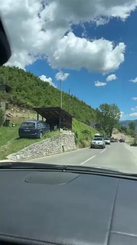 The video shows a view from inside a car driving on a road. The road is paved and appears to be in a rural or mountainous area, with lush green hills on one side and trees lining the other. The sky is blue with fluffy white clouds. Several other vehicles are visible on the road, including a silver SUV with a roof rack, a white van, a black car with a roof rack that appears to be decorated, and a black Volkswagen Golf. Some of the vehicles are driving in the same direction as the camera car, while others are moving in the opposite direction. The scene appears to be set during the daytime, likely in the spring or summer. The overall impression is a scenic drive through a picturesque landscape.