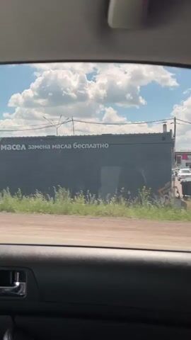 The video shows a view from inside a car looking out the passenger window. The car is moving along a road with a dirt shoulder and some grass and bushes. On the right side of the road, there are several commercial buildings. The first building visible has a dark facade with text that appears to be in Russian, including the words "мир масел" and "замена масла бесплатна". Next, there is a traffic light with a red light illuminated, followed by a building with the text "шинмонтаж". Further along, a building displays the word "АККУМУЛЯТОРЫ". There are also other structures, including what looks like a billboard. The sky is blue with fluffy white clouds. Vehicles are visible on the road, including cars and a truck. The interior of the car is visible at the bottom of the frame, showing a door panel and a handle.