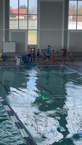 The video shows an indoor swimming pool with two people in the water and a group of children and an adult standing at the edge of the pool. The pool water is mostly still with ripples and reflections. The pool is divided into lanes with black and white lane markers. A green and yellow floatation device is floating in the middle of the pool. A metal bar is stretched across the pool between the two lanes. At the side of the pool are a number of children in swimming gear, including caps. A man in a blue shirt is standing with them. A blue slide is next to them, with a child sitting at the top. One person in the pool is swimming, and another is standing with their back to the camera, holding on to the pool ladder. Large windows are visible in the background.
