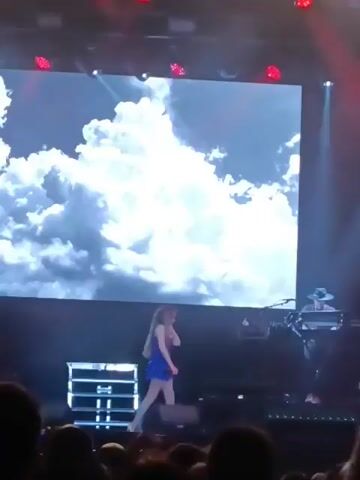 The video shows a live stage performance. A young woman with long hair, wearing a sparkly blue dress, walks across the stage. Behind her, a large screen displays a cloud image. A man in a hat plays a keyboard to her right. As she gets closer to the right side of the stage, a man in a dark hoodie and jeans approaches her, and they briefly touch hands, possibly a high-five or a gesture of support.  There is a stage prop of a floral frame in the background. Stage lighting is prominent, with red and white beams illuminating the scene. A crowd is visible in the foreground, with a phone recording the event.