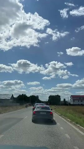 A car is driving down a road with traffic. The sky is blue with white clouds. There are houses and trees on the side of the road.