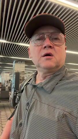 A man wearing glasses and a baseball cap is in an airport. He is wearing a grey checkered shirt. He is looking at the camera and talking. Behind him are rows of airport seats and a departure board. The airport appears to be modern with a ribbed ceiling and fluorescent lighting.