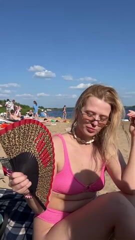 A woman with blonde hair and sunglasses sits on a sandy beach. She wears a pink bikini and a seashell necklace. In her right hand, she holds a black folding fan with a leopard print and red accents. The fan is partially open and angled towards her face. Behind her, other beachgoers are visible, some lying on towels and others standing. The sky is blue with scattered clouds, and a body of water is in the background. The woman is fanning herself with the fan.