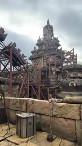 The video shows a rollercoaster moving quickly across a track system built around a tall, weathered stone structure. The structure resembles an ancient temple or fortress, with multiple levels and intricate carvings. The rollercoaster tracks are supported by a red metal framework, adding to the scene's industrial aesthetic. In the foreground, there is a stone wall, a wooden chest, and a coiled rope. The sky is overcast, suggesting a cloudy day.