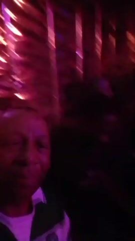 The video depicts a man taking a selfie video in a dimly lit nightclub or bar with red and blue lighting. Other people are visible in the background, sitting at a table. A sign with the word "KASK" is visible on the wall behind them. Loud music can be heard in the background.