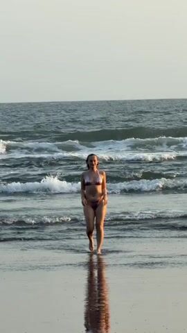 A woman in a bikini walks along a wet sandy beach toward the camera. The ocean is behind her with waves breaking and rolling towards the shore. The sky is a pale, hazy color. The woman is smiling and looking up and to her right. Her reflection is visible in the wet sand.