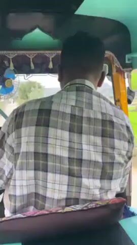 From inside a yellow and teal auto-rickshaw, the video shows the driver's back, wearing a plaid shirt, as the vehicle drives past bright green rice paddies under a cloudy sky. The backdrop is a landscape featuring rocky hills and scattered trees, with utility poles and wires visible in the distance.